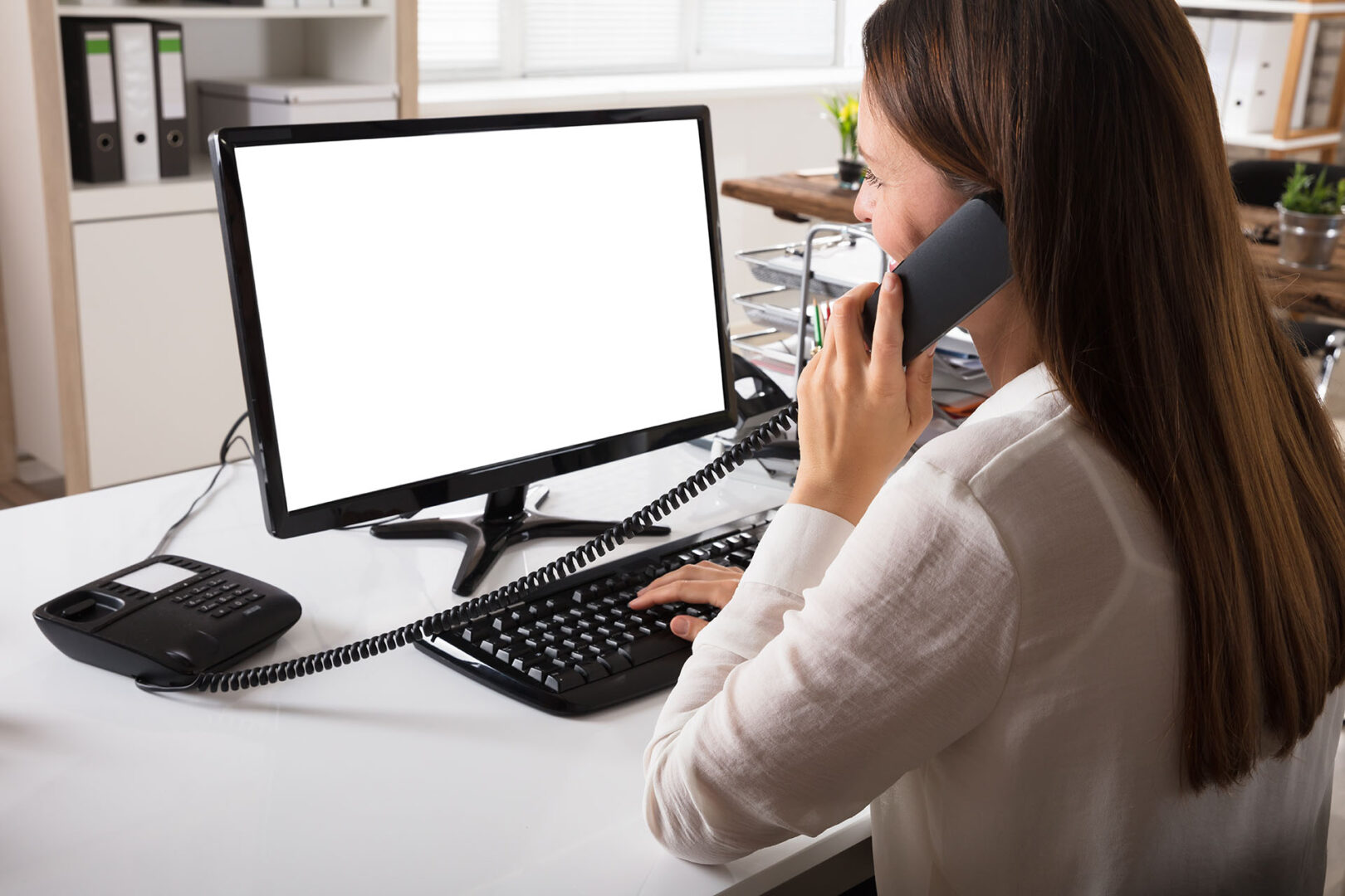 Businesswoman Talking On Landline Phone With Blank White Computer Screen On Desk